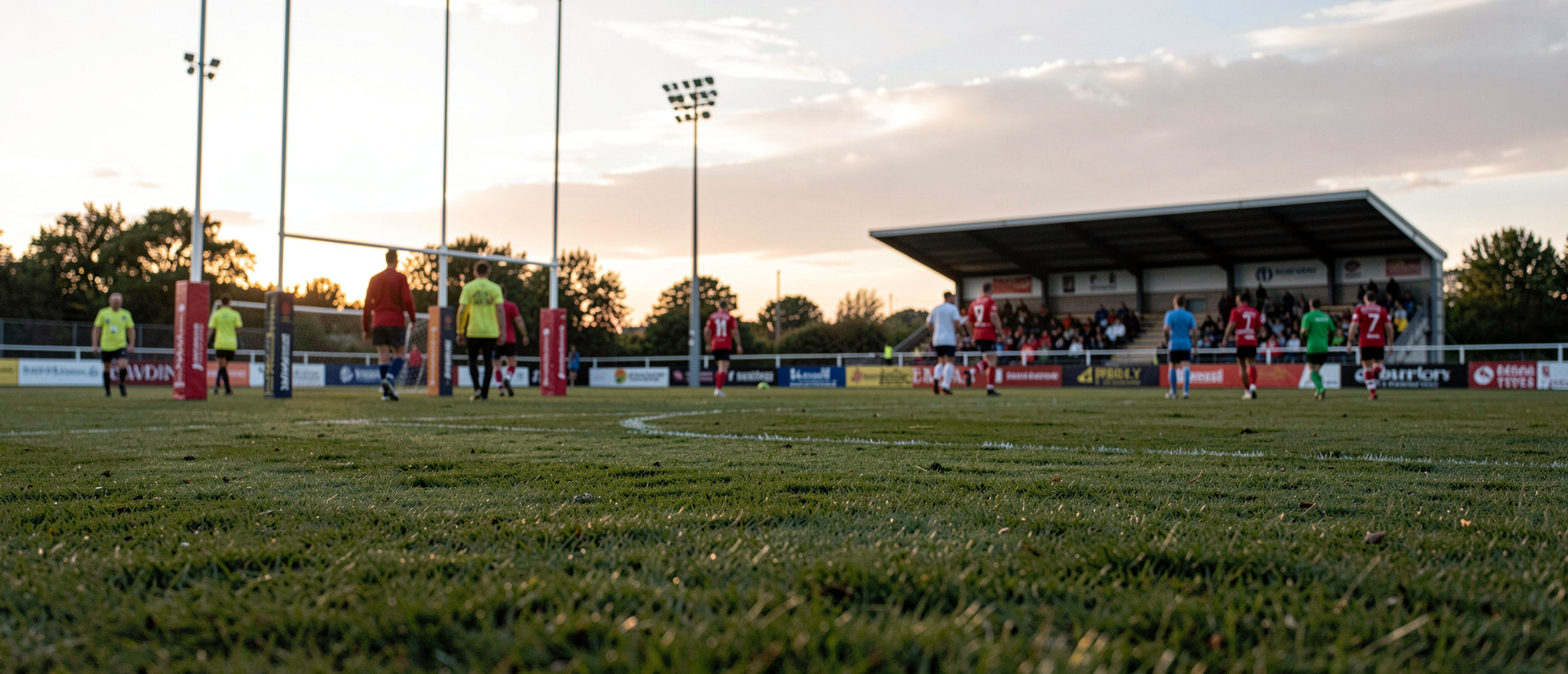 Kvällsljus över en rugbyplan med potealen resta mot gräset, åskådare på väg mot en av de fyra tribUNERNA, kameran i marknivå från sidlinjen