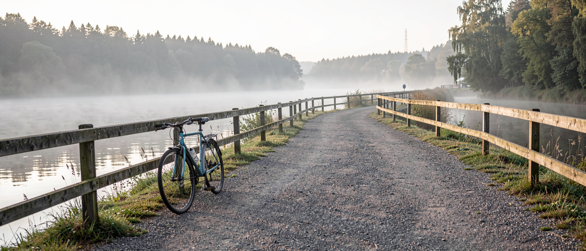En flack grusväg längs floden Fulda en tidig söndagsmorgon, låg dimma över vattnet, en cykel lutad mot ett träräcke i förgrunden och skogsbryn i bakgrunden, fotograferat i ögonhöjd med mjukt sidoljus.